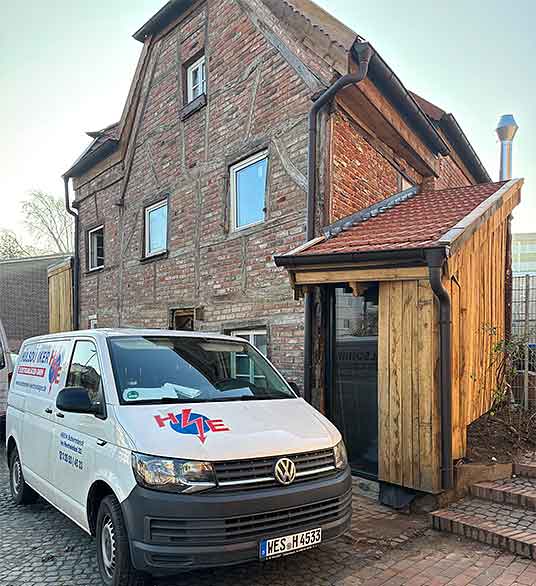Weißer Volkswagen Transporter mit HSE-Logo steht vor einem alten Fachwerkhaus mit rotem Ziegeldach und Holzverkleidung an der Eingangsseite.