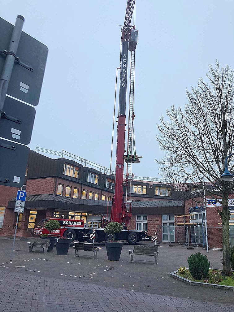 Ein roter Kran mit der Aufschrift 'SCHARES' hebt eine Last vor einem Backsteingebäude auf einem Platz mit Bänken und einem Baum.