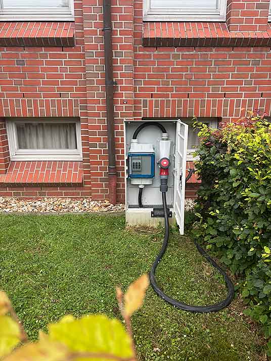 Offener Stromverteilerkasten an einer Backsteinwand mit einem angesteckten schwarzen Kabel auf einem Rasen neben einem Busch.