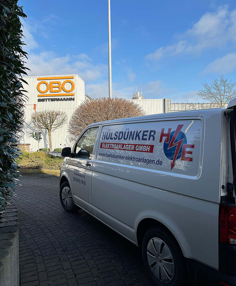 Weiße Firmenfahrzeug mit Werbung von Hülsdünker Elektroanlagen vor dem OBO Bettermann Gebäude bei sonnigem Himmel.