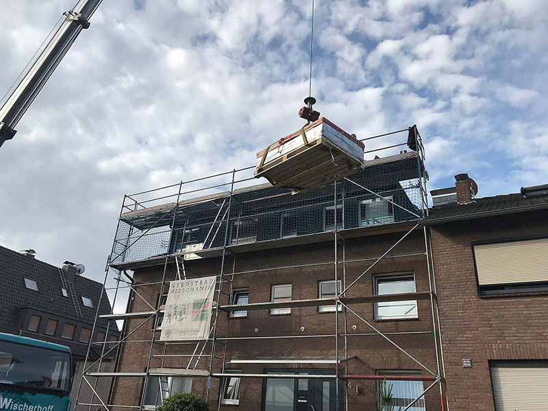 Kranausleger hebt eine Palette mit Baumaterialien über ein eingerüstetes zweistöckiges Wohnhaus.