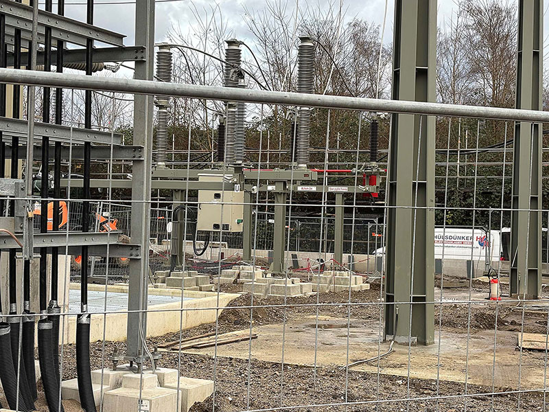 Baustelle eines Umspannwerks mit Stahlkonstruktionen, Isolatoren und Kabeln hinter einem Drahtzaun.