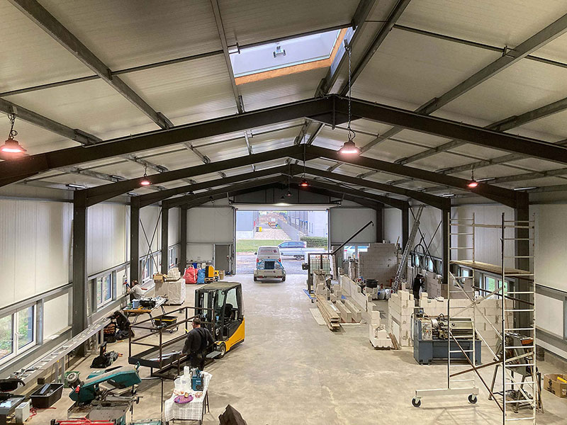 Innenansicht einer Werkhalle mit einem gelben Stapler, Baumaterialien und Regalen, durch die offene Torwand ist ein kleiner weißer Lieferwagen draußen zu sehen.